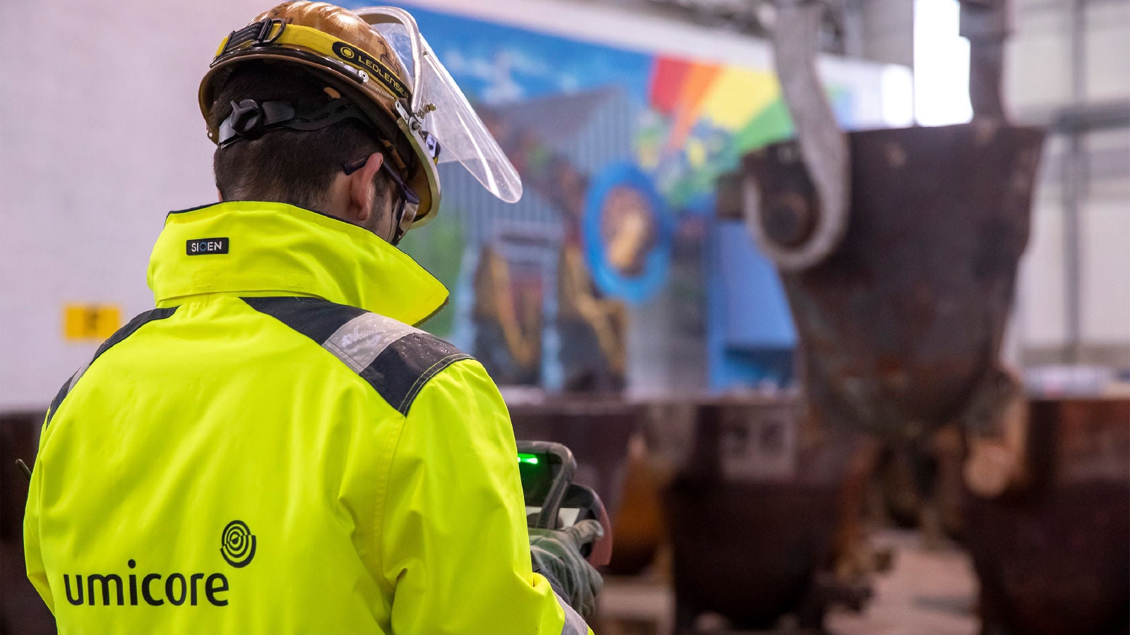 A worker in a high-visibility yellow jacket and hard hat operates a handheld device inside an industrial facility, with large metal containers and a colorful mural visible in the background.