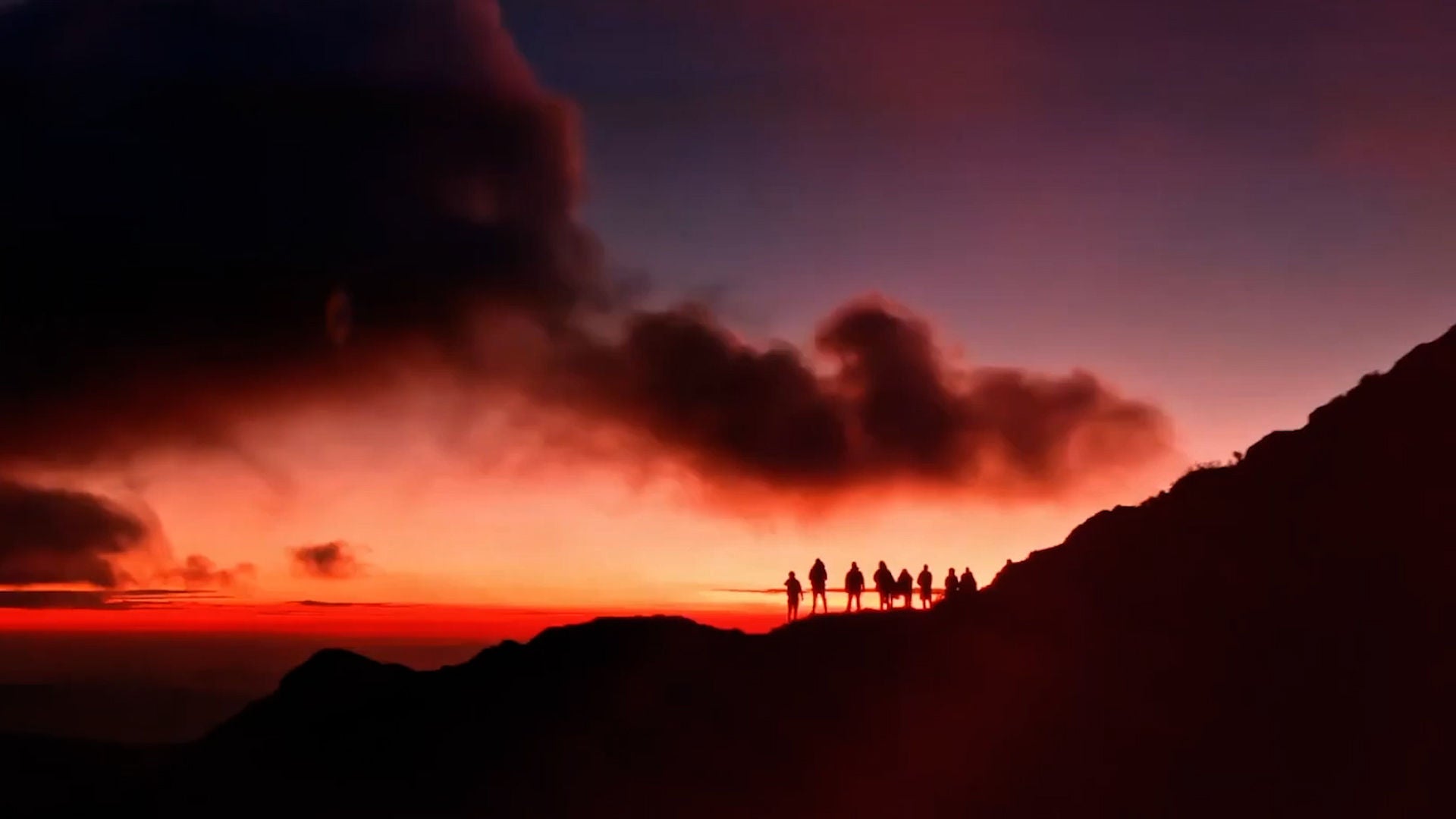 Silhouettes of people stand on a mountain ridge against a colorful sunset sky.
