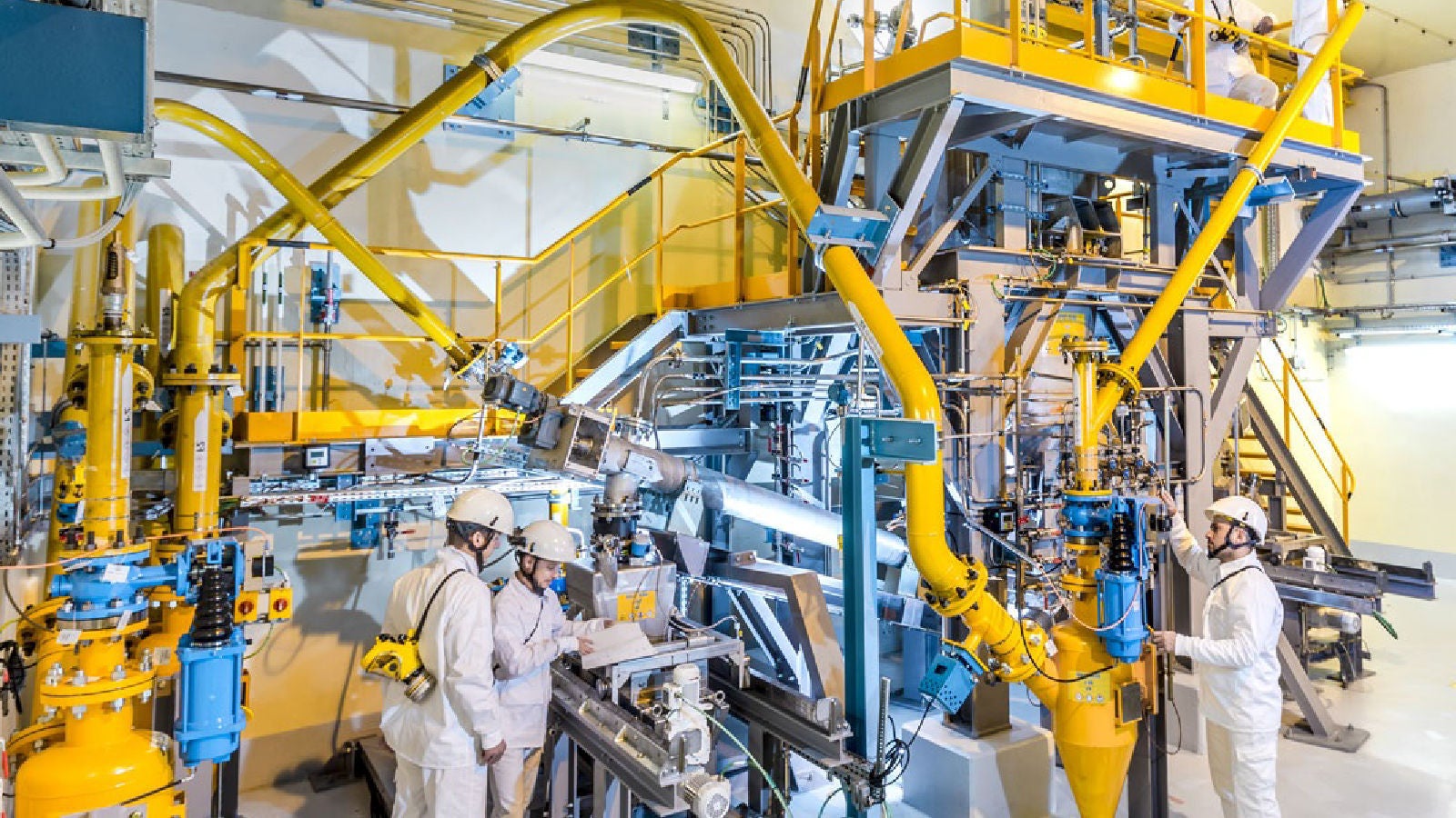 Wide view of a brightly lit, complex industrial plant interior with multiple workers in white safety suits and helmets operating machinery and reviewing documents.