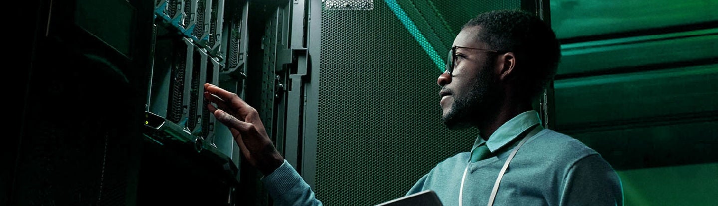 A professional in a data center adjusting server equipment while holding a laptop. The scene is illuminated with green lighting, giving a high-tech ambiance.