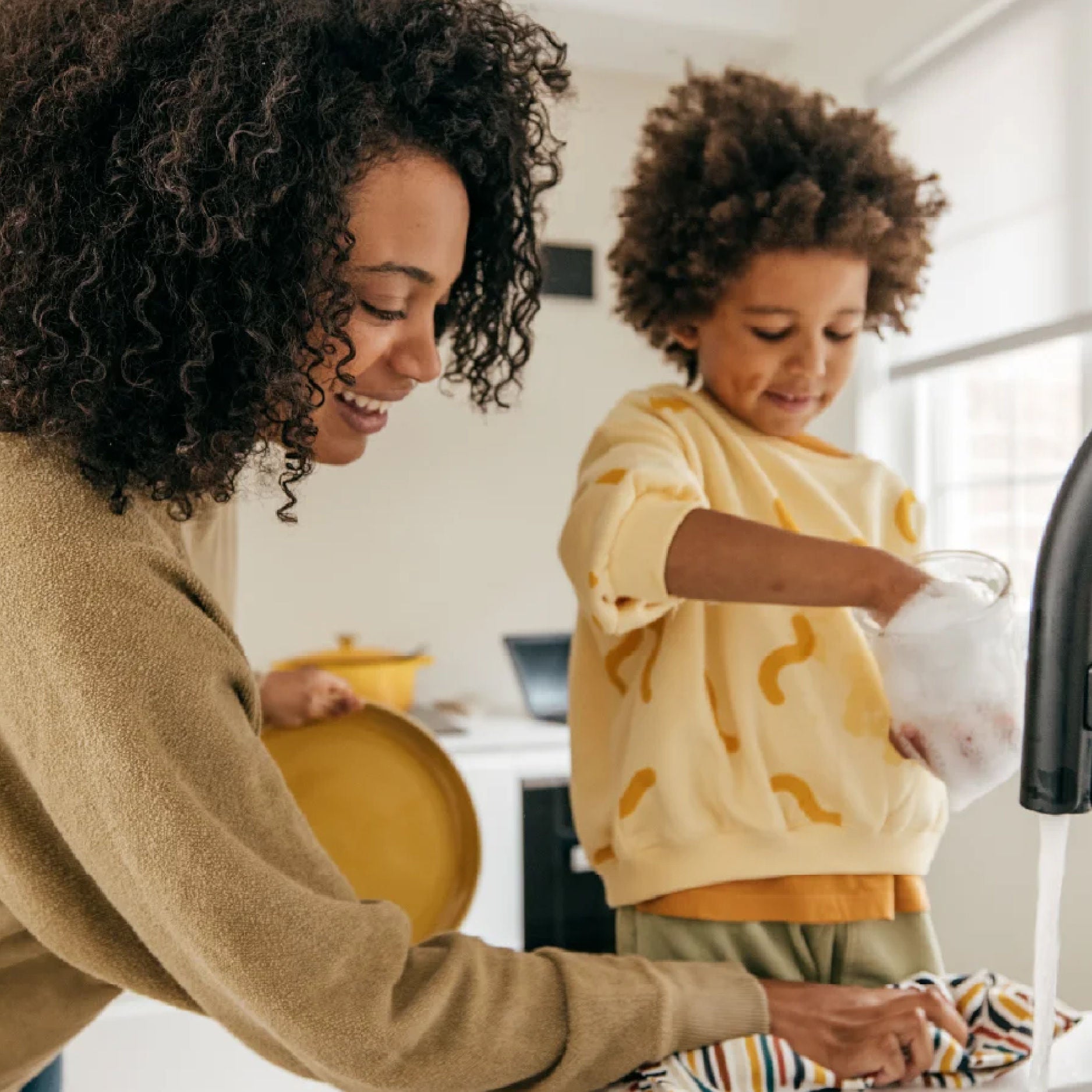 Parent and child preparing food together in a bright kitchen, with the child pouring ingredients while standing beside a running sink.