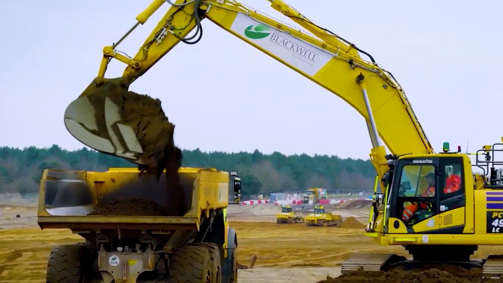 A yellow excavator labeled “Blackwell Earthmoving” dumps soil into a large dump truck at a construction site with other machinery in the background.