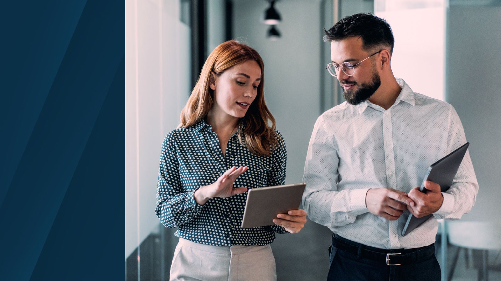 Two professionals walking through a modern office hallway, reviewing data on tablets while engaged in discussion.