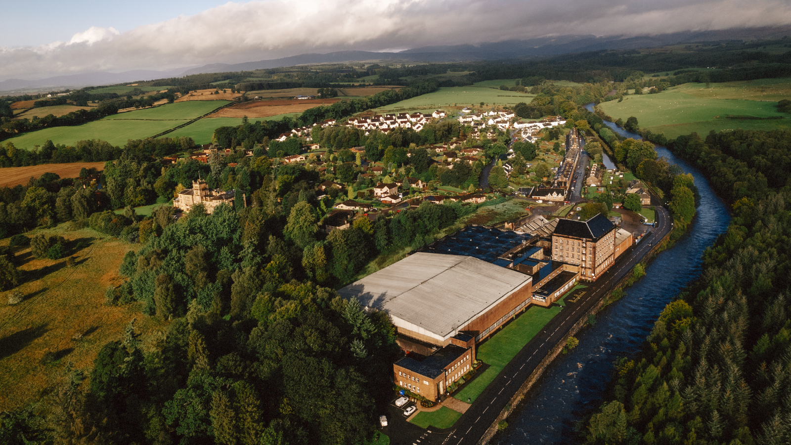 A scenic aerial view of a countryside distillery surrounded by green fields, a river, and a small village.