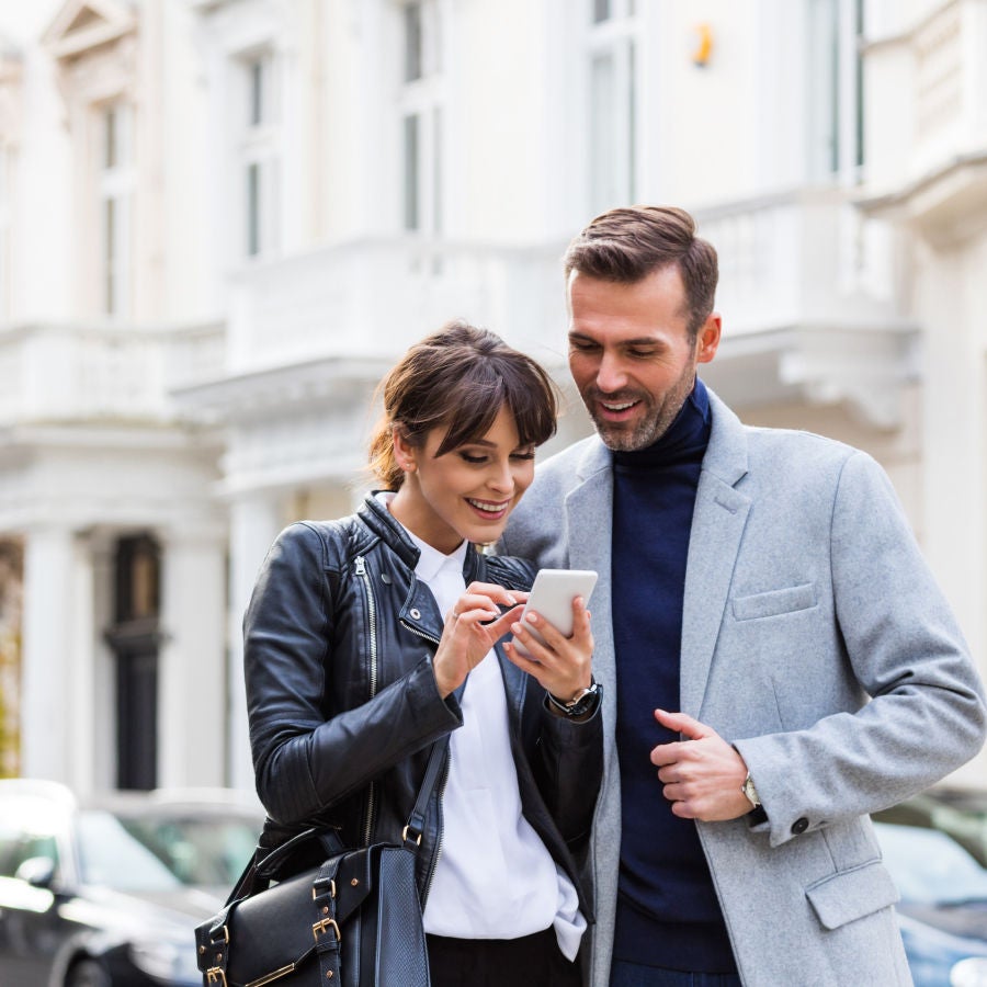 A man and woman stand before a building, both focused on a cell phone in their hands, engaged in conversation.