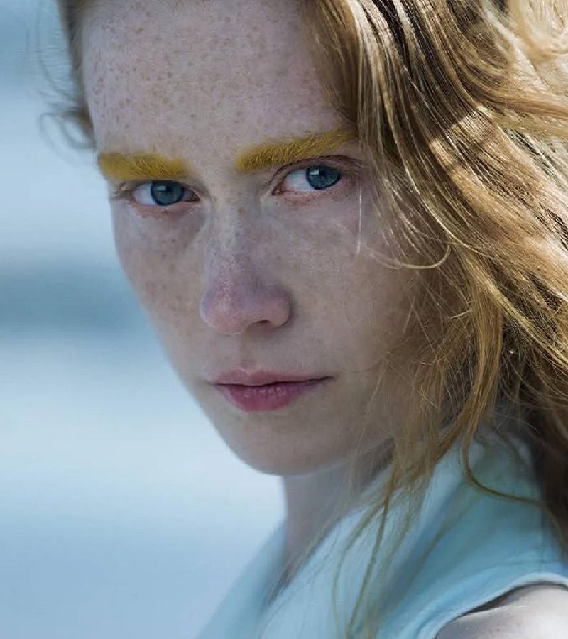 Close-up portrait of a woman with striking yellow eyebrows and intense blue eyes, set against a soft outdoor background.