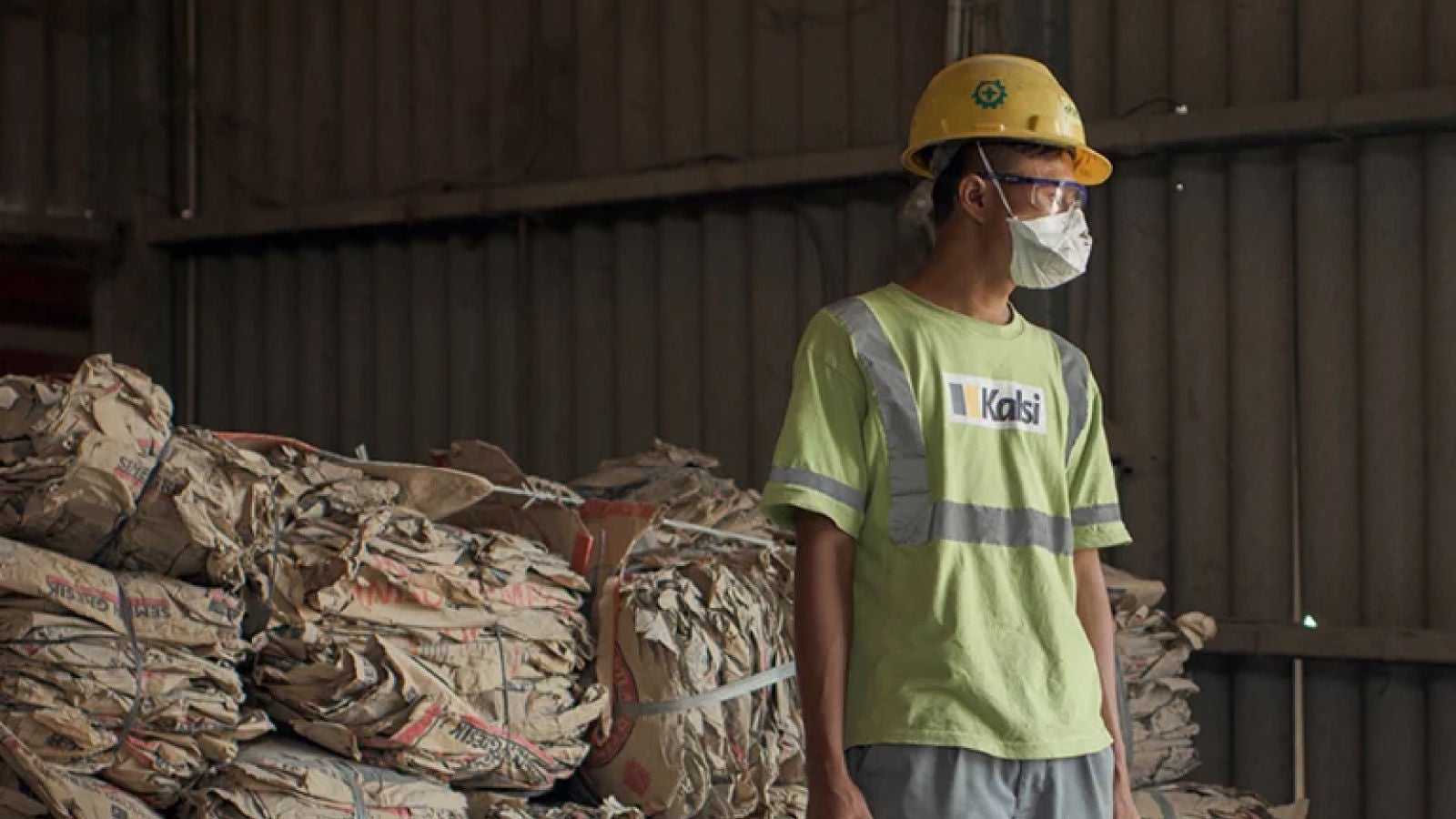 A worker in a green shirt and yellow hard hat stands in a warehouse with stacks of compressed paper packaging behind him. He wears a face mask and safety goggles.