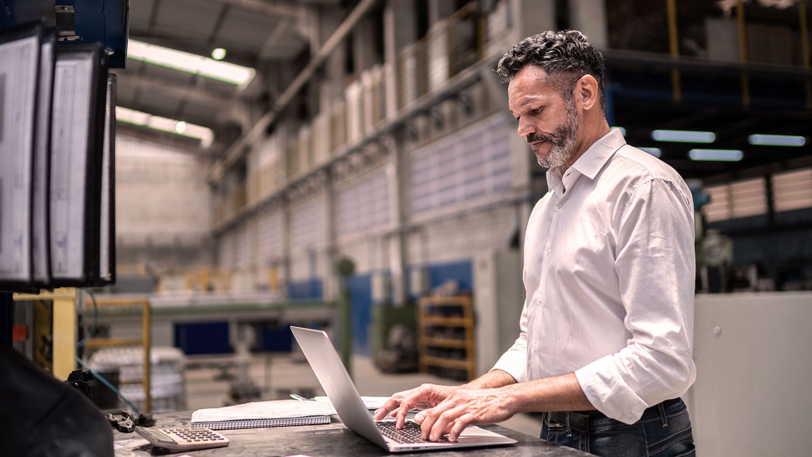 A manufacturing executive in a factory setting, working on a laptop with industrial equipment and documents in the background, representing data-driven decision-making in consumer products planning.