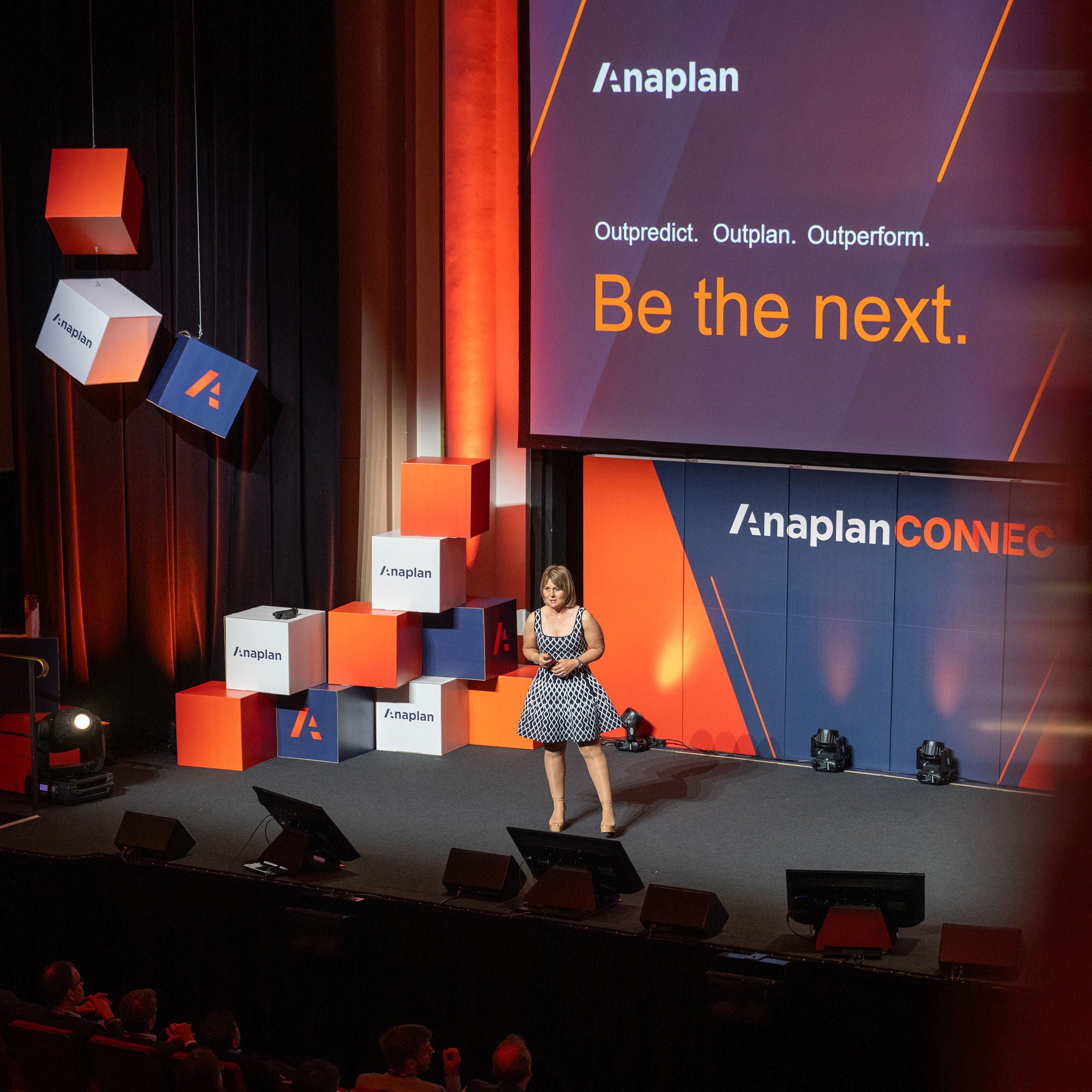 Anaplan Connect event photo. A speaker stands on stage in front of a large screen displaying the text “Outpredict. Outplan. Outperform. Be the next.” with Anaplan branding. The stage is decorated with stacked and hanging branded cubes in orange, navy, and white.