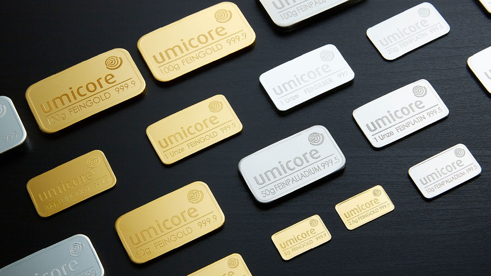 A variety of gold, silver, platinum, and palladium bars branded with the Umicore logo are arranged on a black surface, showcasing different weights and metal types.