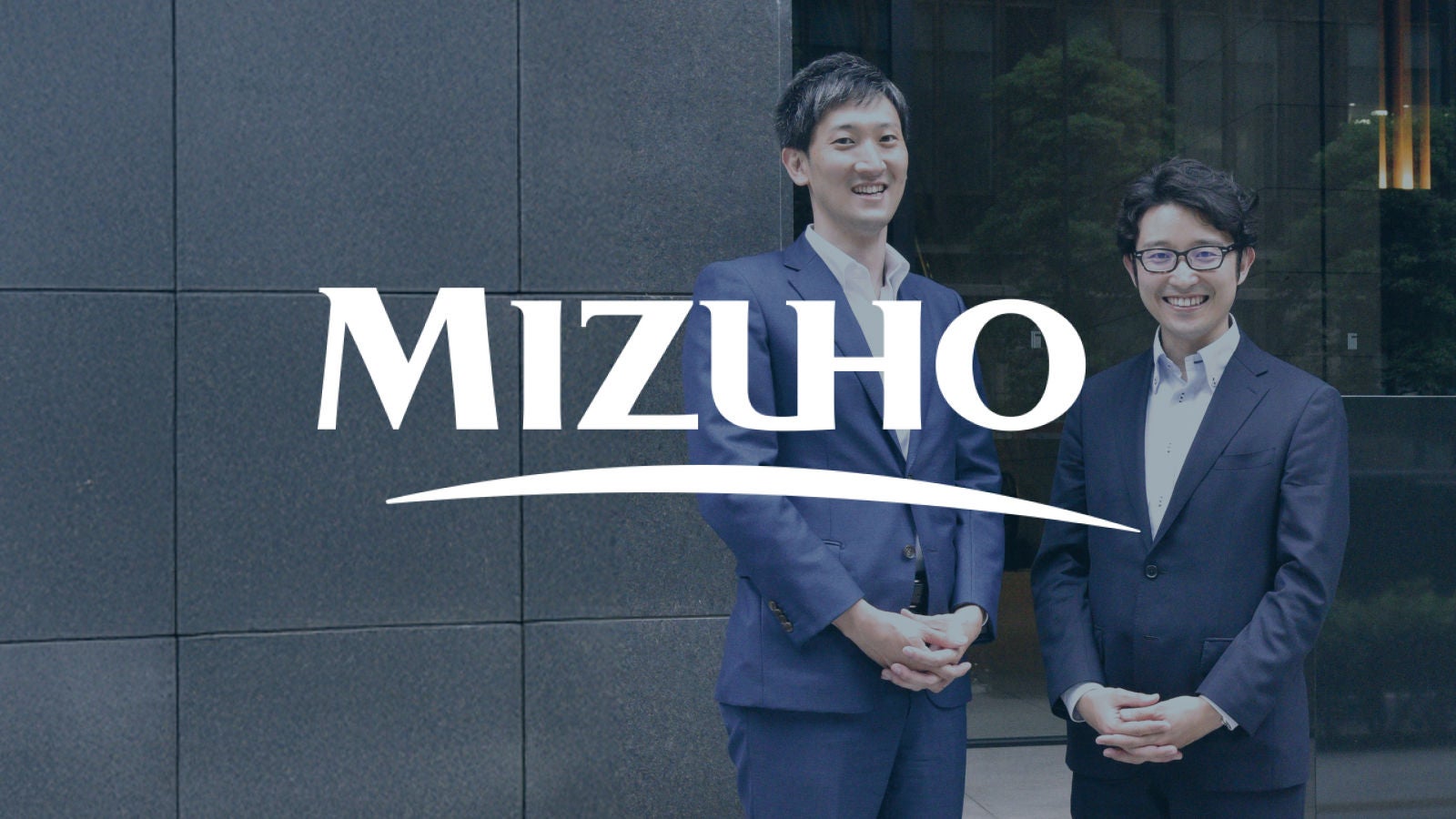 Mizuho logo displayed over a background showing two men in business suits standing outside a modern office building.