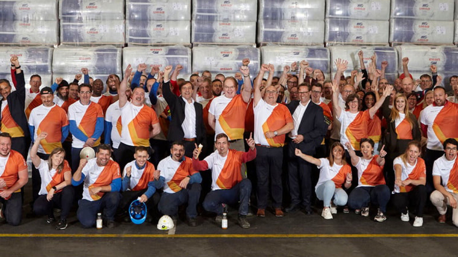 A large group of employees wearing matching orange and white shirts celebrate together in a warehouse, raising their hands in excitement.