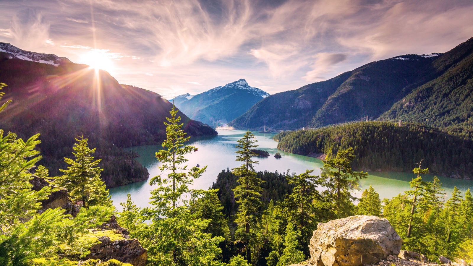 Scenic view of a mountain landscape with evergreen trees, a calm lake, and sunlight streaming through the clouds.