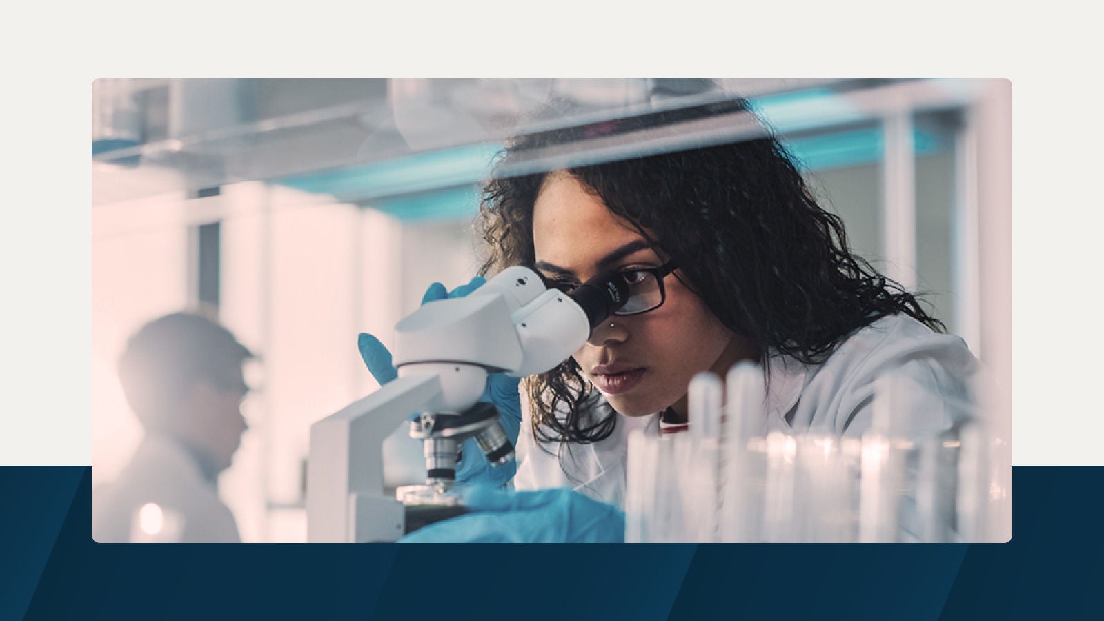 A scientist wearing gloves and glasses looks through a microscope in a laboratory, surrounded by test tubes and research equipment.
