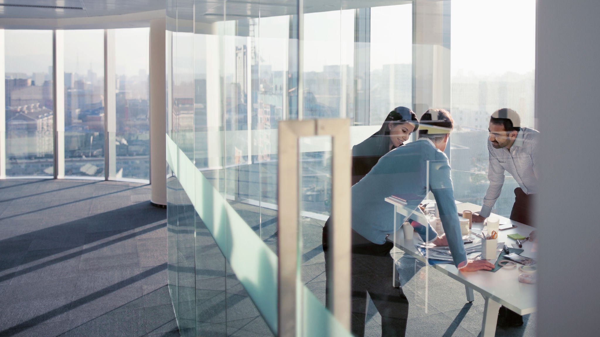 Three employees having a meeting in a glass-walled conference room in a high-rise building.