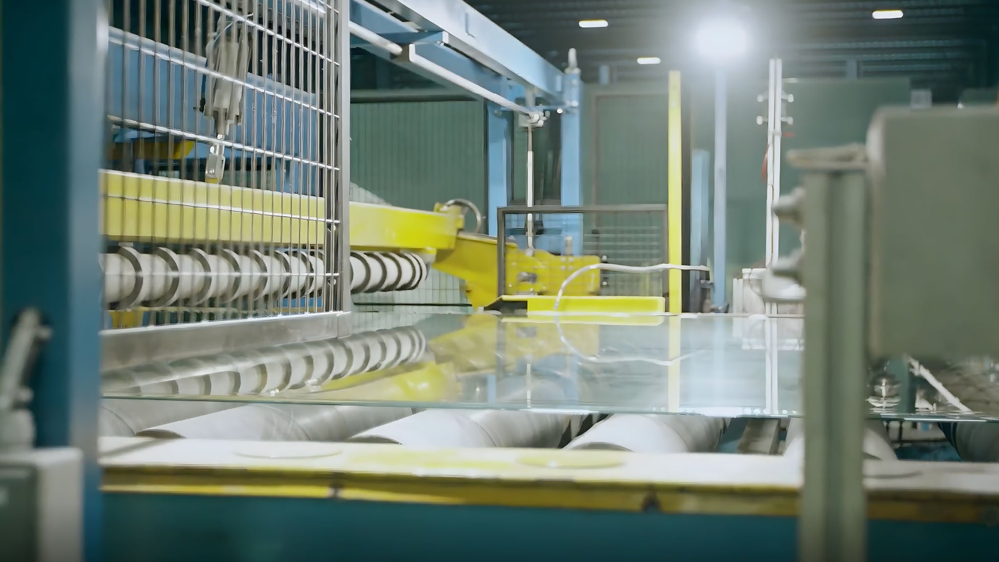 Factory scene with yellow and blue conveyor machinery transporting clear glass sheets through a production line.