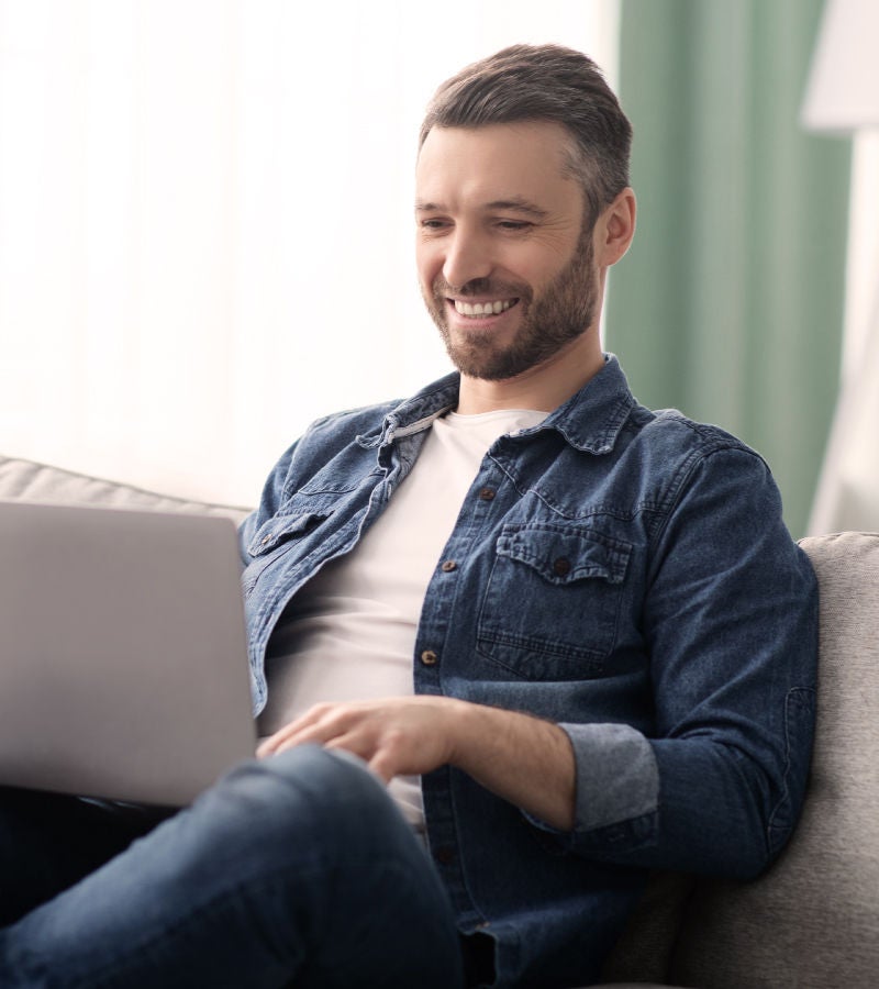 Smiling man in a denim shirt working on a laptop while sitting comfortably on a couch in a bright living room.