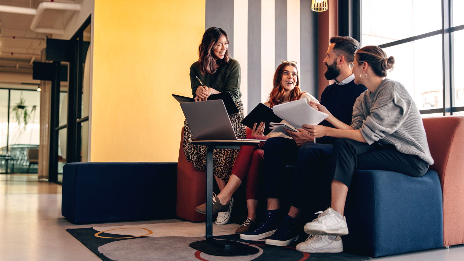 Group of four young professionals sitting in a modern office lounge, smiling and collaborating with notebooks and a laptop in front of a striped wall and large windows.