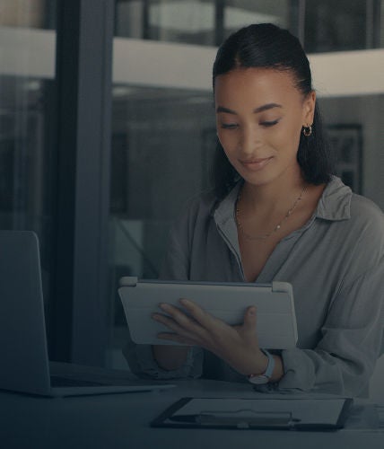 Woman using a tablet at her desk, with a laptop and notepad beside her, smiling while working.
