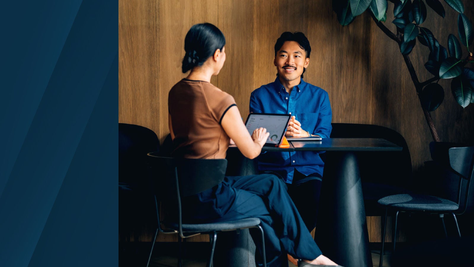 The image shows a closer view of a woman using a tablet while speaking to a smiling man seated across from her at a small round table in a warm-toned office space.