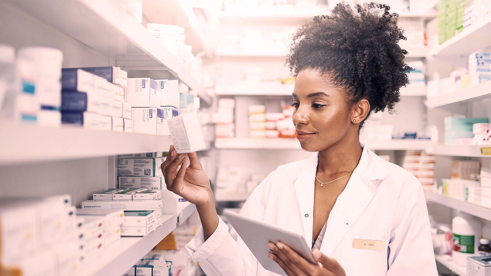 Pharmacist in a white coat examining a medicine box while holding a tablet, surrounded by shelves filled with pharmaceutical products.