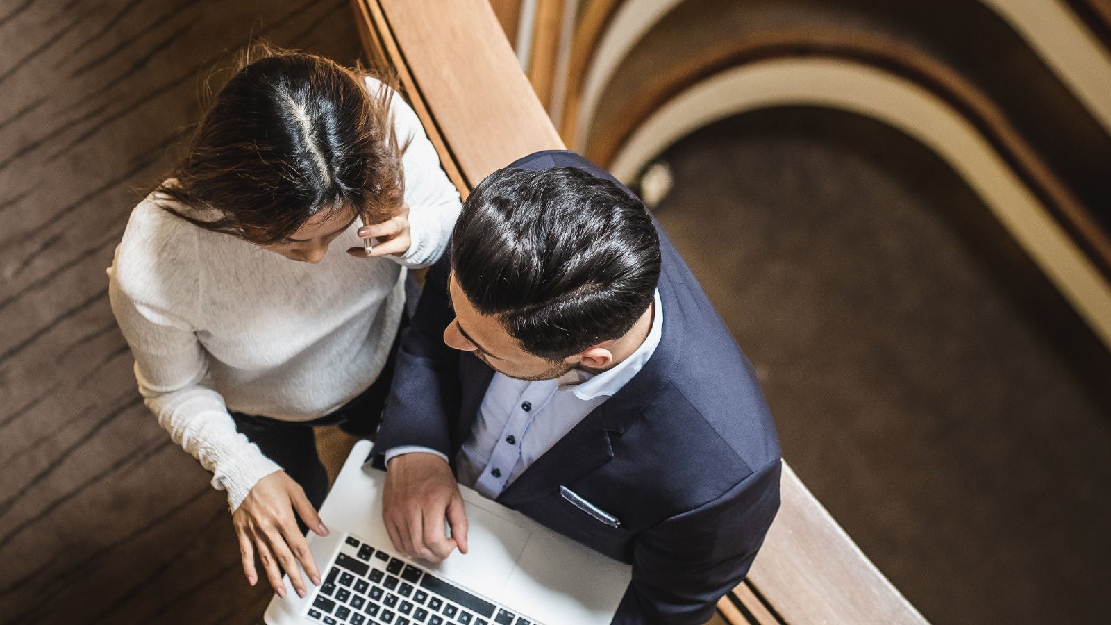Top-down view of a man in a suit and a woman in a white sweater standing by a staircase, looking at a laptop and discussing business strategy.
