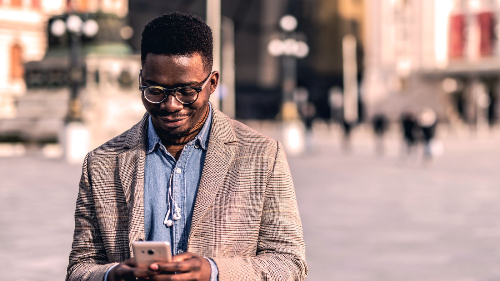 A man in a checkered blazer and glasses smiles while looking at his smartphone outdoors in a city square.
