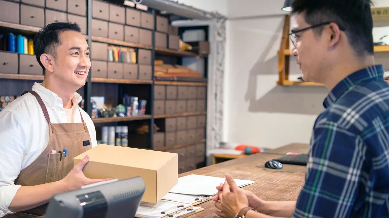 A smiling store worker wearing an apron hands a package to a customer across a wooden counter in a shop with neatly organized drawers.