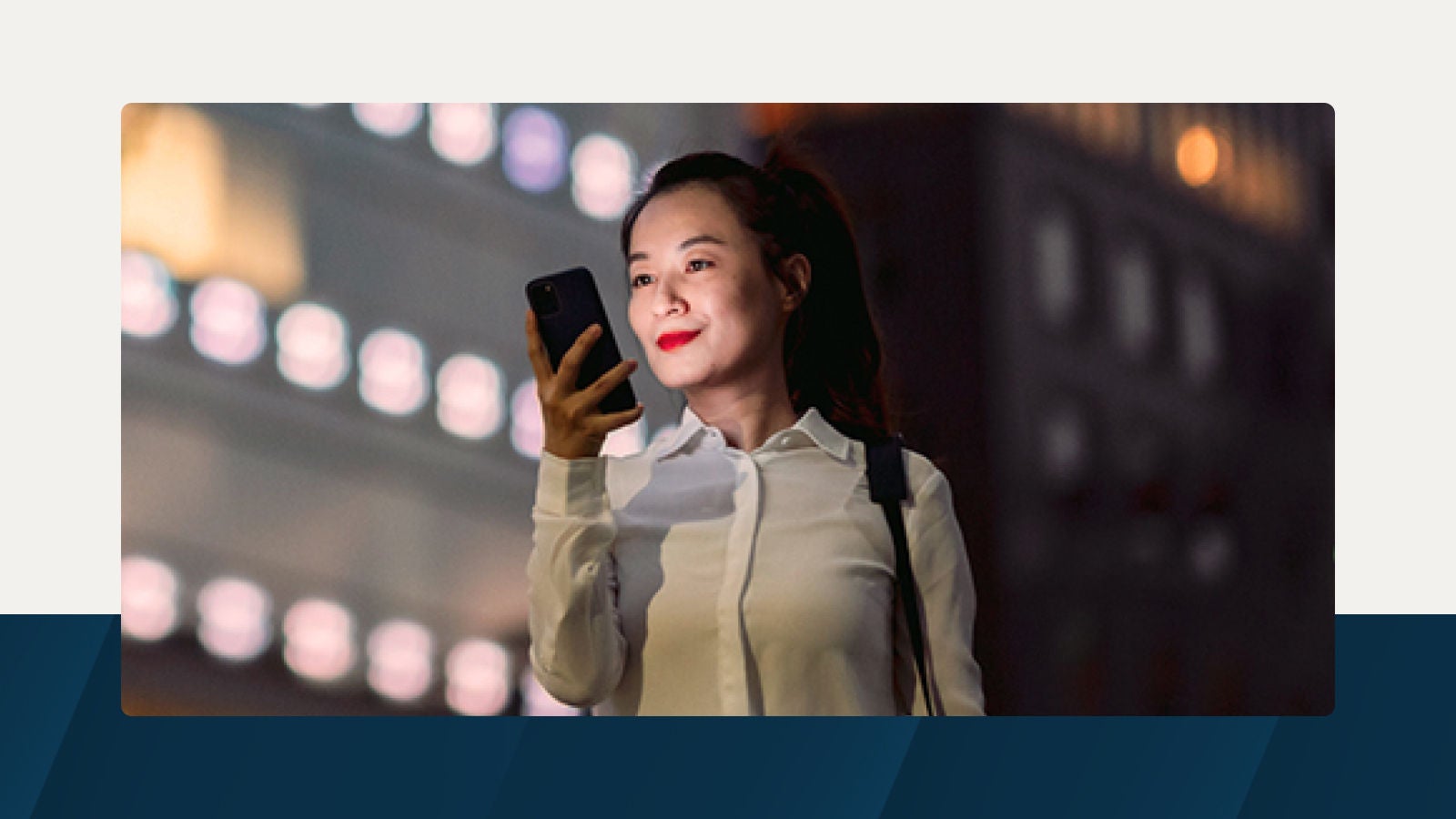 A professional woman in business attire smiles while looking at her phone at night, illuminated by city lights, symbolizing digital transformation in capital markets.