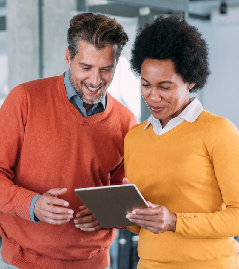 Two professionals in a modern office setting, smiling and collaborating while using a tablet.