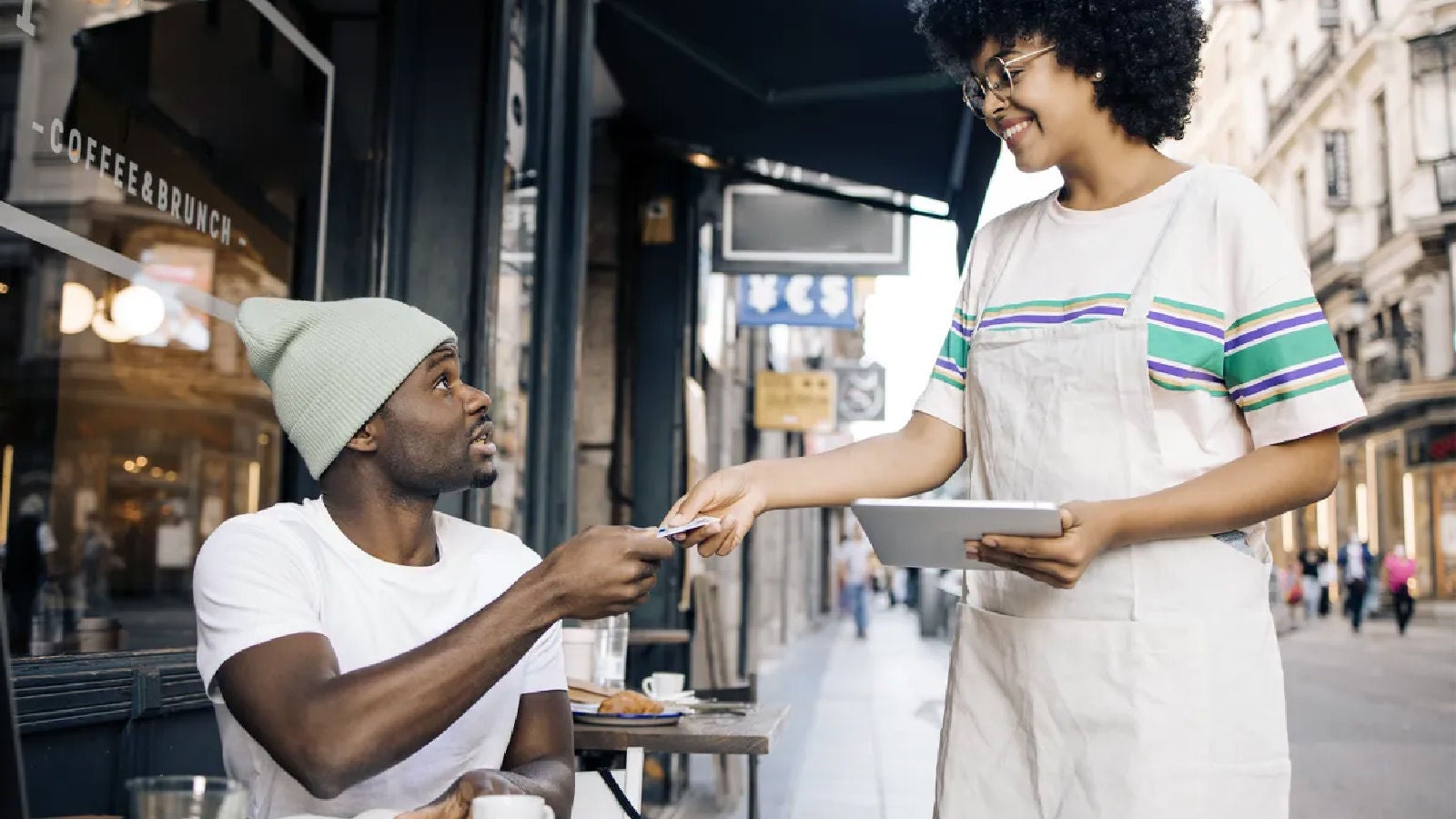 A smiling café worker in an apron accepts a card payment from a seated customer outside a street café.