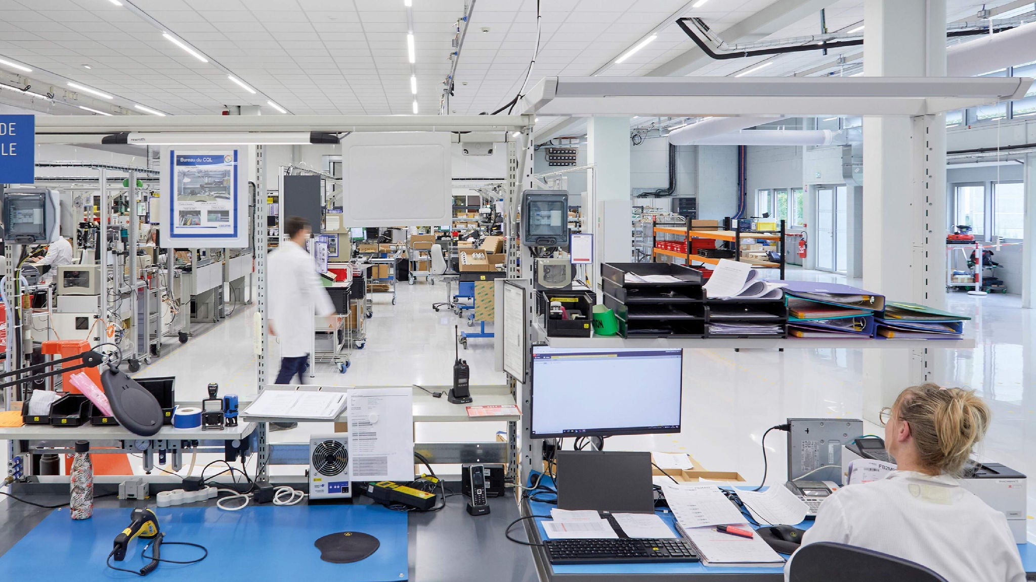 Modern manufacturing facility with employees in lab coats working at organized desks and assembly stations.