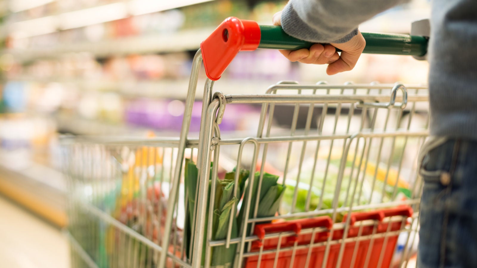 Close-up of a person pushing a shopping cart filled with groceries in a brightly lit supermarket aisle.