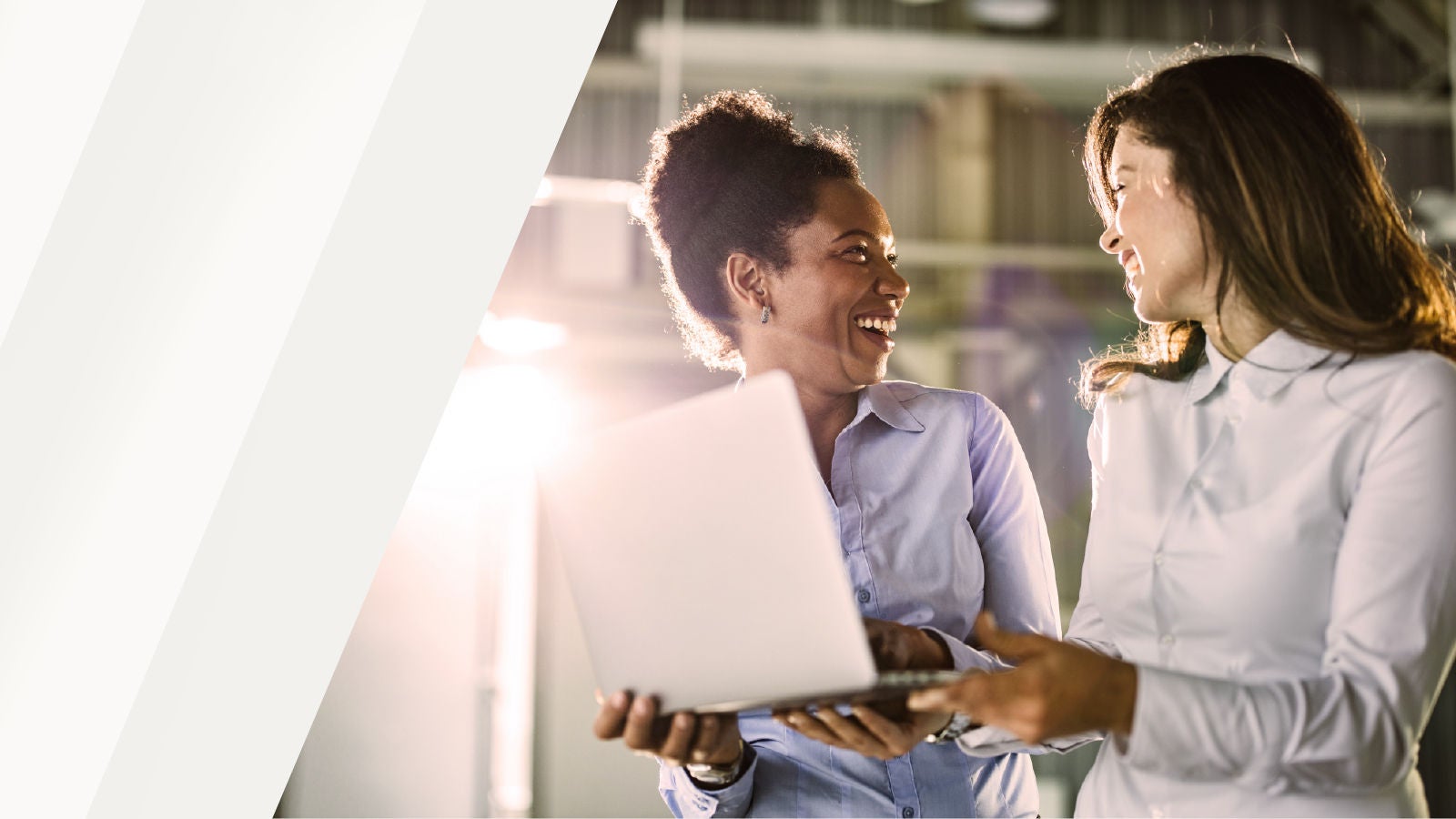 Two professionals smiling and talking while holding a laptop, with sunlight shining through an industrial office space.