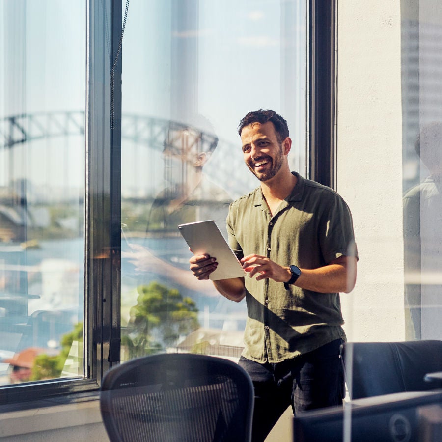 A smiling man engaged with a tablet while seated at a desk in a modern office environment.