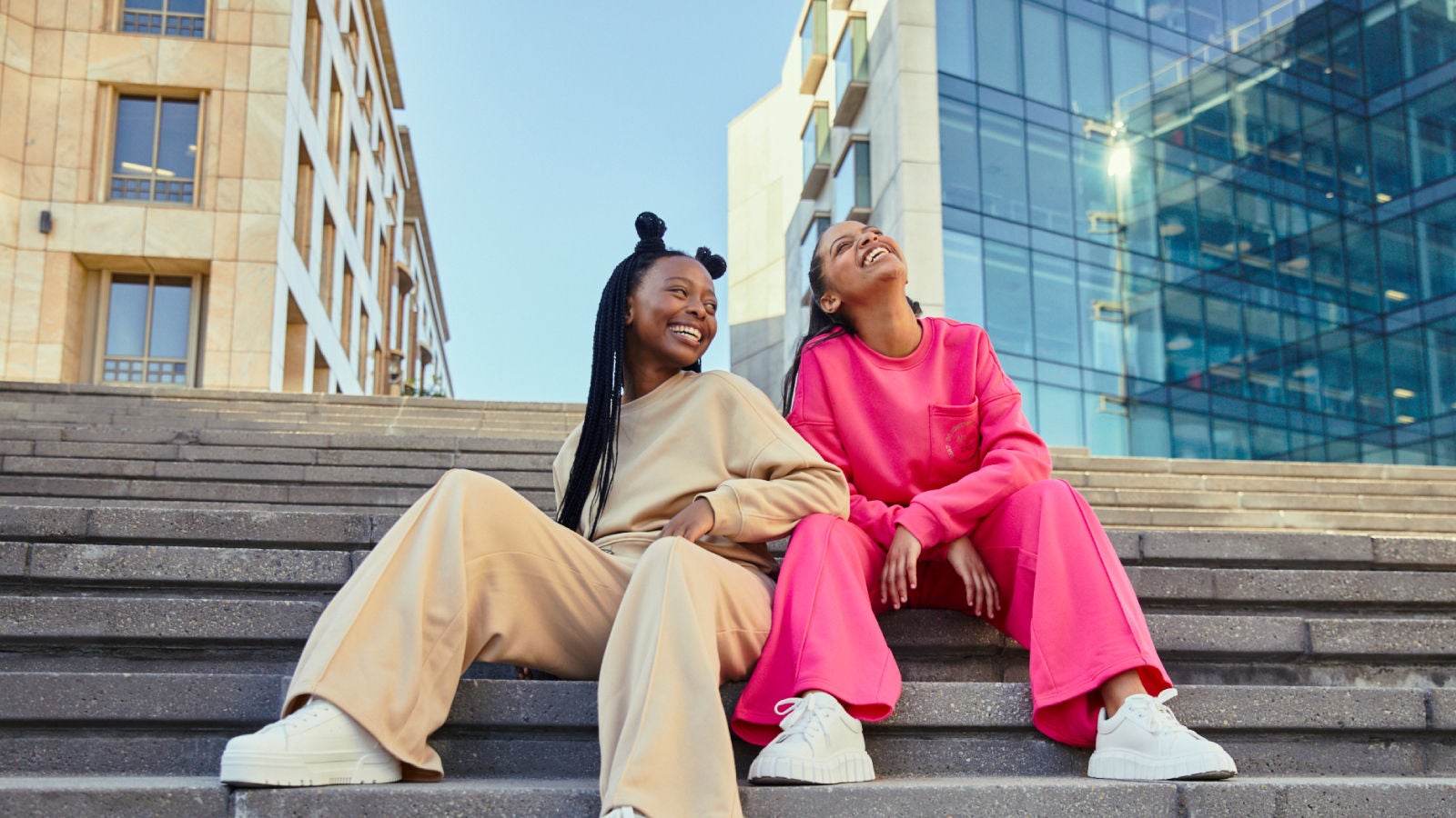 Two women sitting on outdoor steps, dressed in stylish beige and bright pink casual outfits, laughing and looking up, with modern glass and stone buildings in the background.