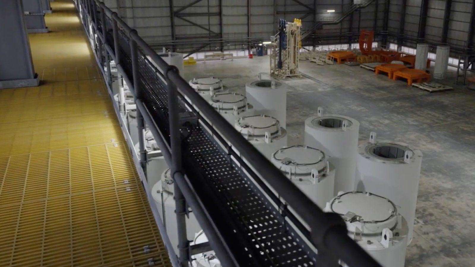 An overhead view of an industrial facility interior with large cylindrical components, metal walkways, and equipment spread across a spacious warehouse.