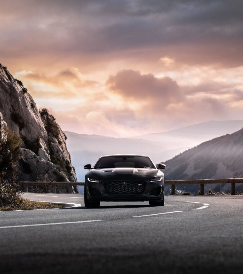A sleek black car driving along a winding mountain road during sunset, with dramatic clouds and rocky cliffs.