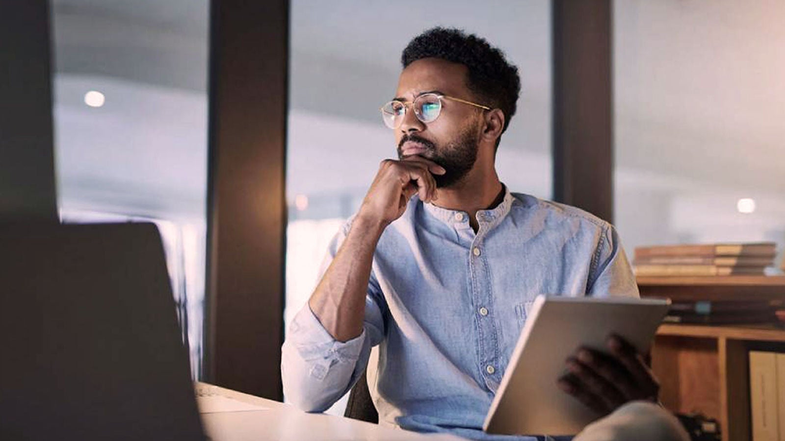 A business professional sits at a desk, holding a tablet, deep in thought, with a blurred background featuring office elements.