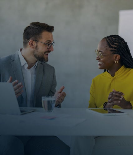 Two professionals seated at a desk, engaged in a discussion with a laptop and glass of water on the table.