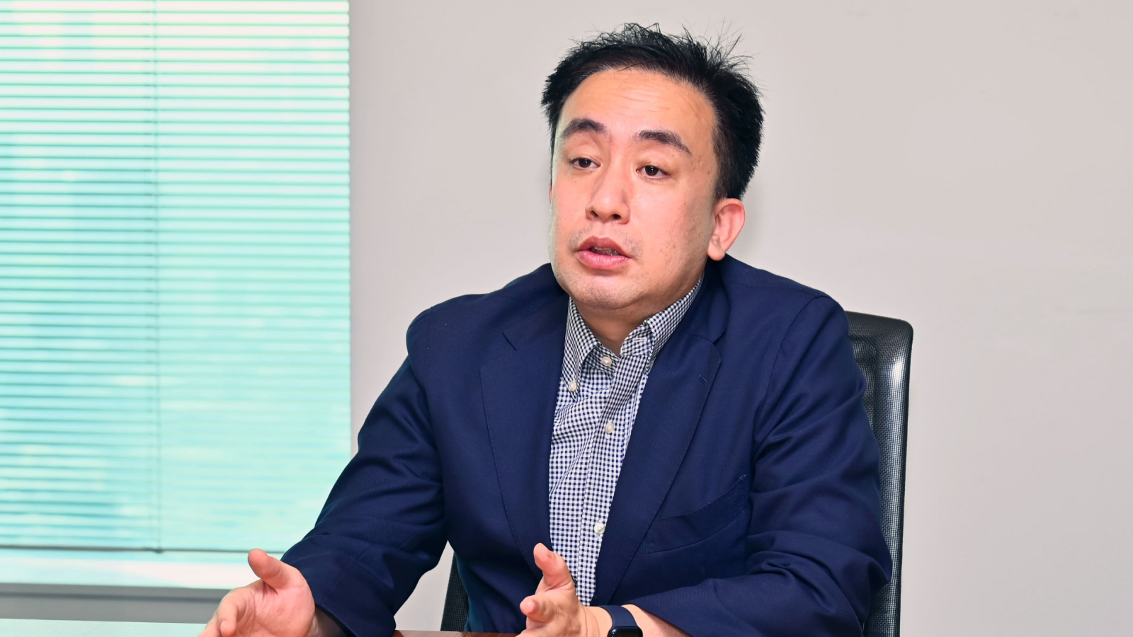 Godiva Japan Customer Story Illustrative Image 1 A man in a navy blazer and checkered shirt sits at a desk, gesturing as he speaks. A window with closed blinds is in the background.