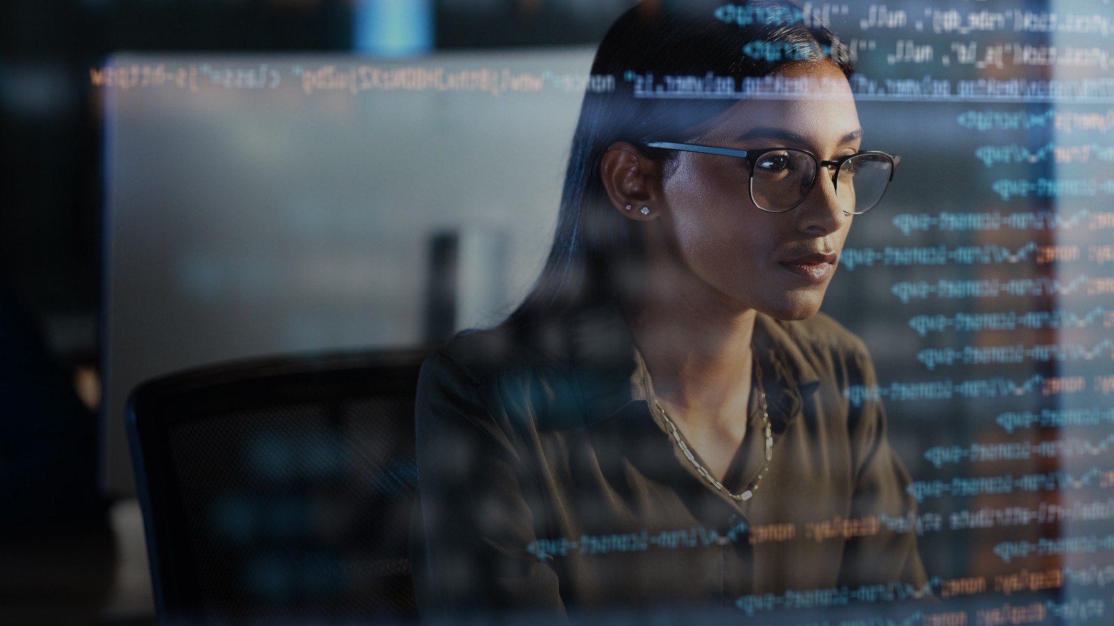 A woman wearing glasses looking at lines of code reflected on a glass surface in a dark, modern workspace.