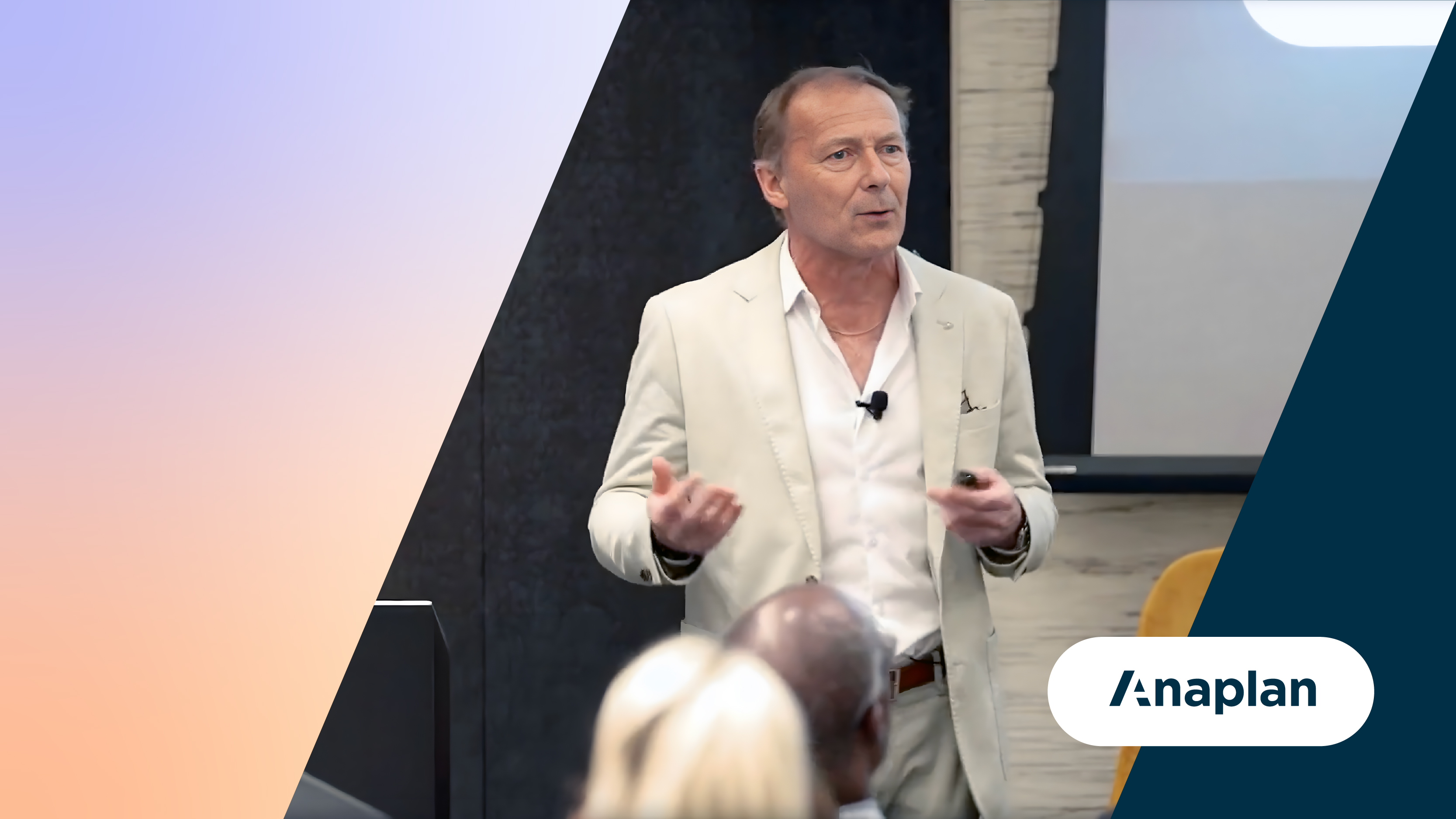 Anaplan Logo displayed bottom right. A speaker in a cream suit gestures while speaking to an audience at an Anaplan event.