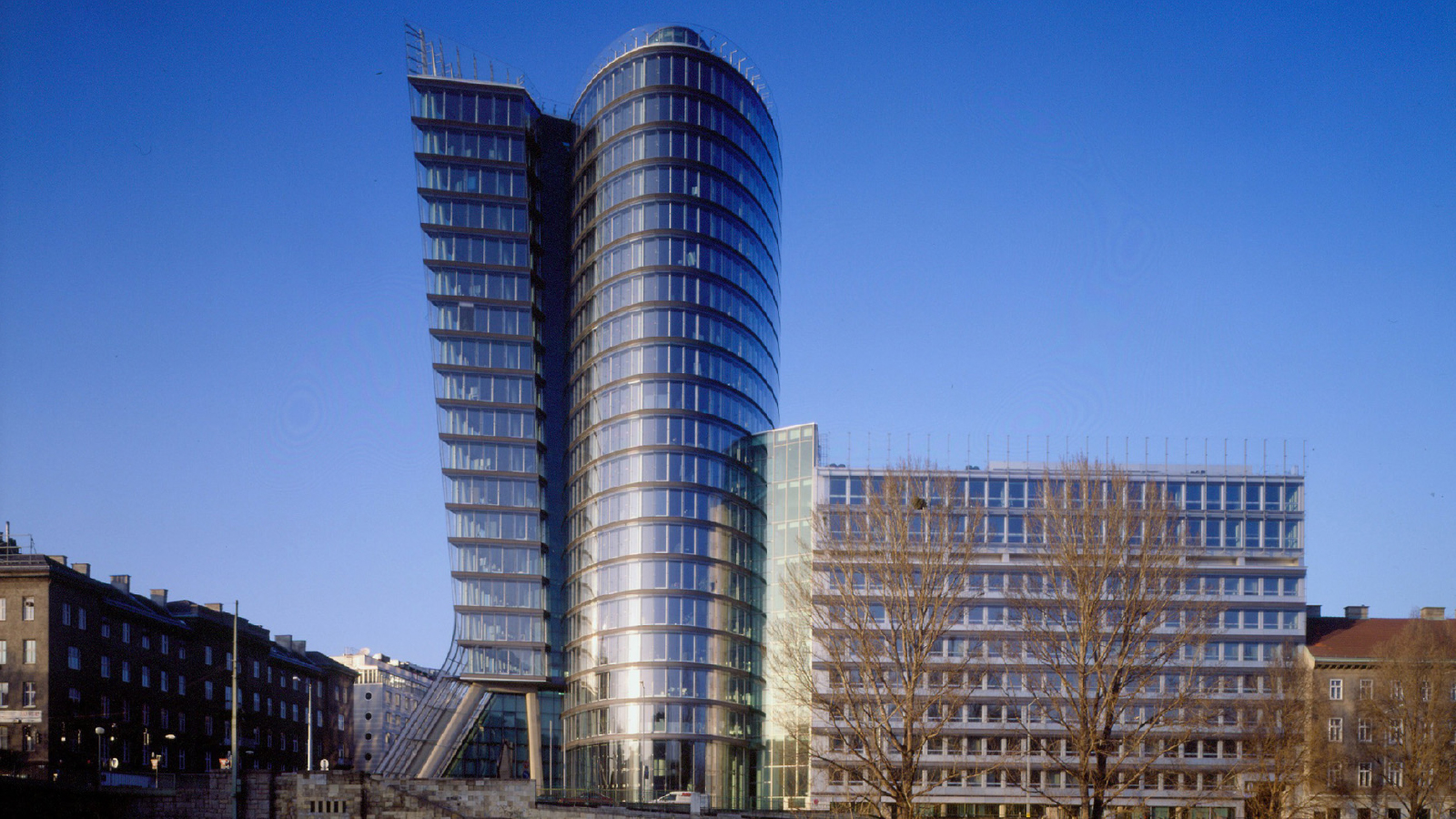 Tall building made of glass walls standing within a city with other shorter buildings surrounding